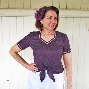 K&ouml;nnte beinhalten: Ein Oberteil mit wei&szlig;en und violetten Punkten und einem rosa Besatz an Ausschnitt und &Auml;rmeln. Das Oberteil ist in der Taille mit einer Schleife gebunden. Das Model tr&auml;gt einen wei&szlig;en Rock.