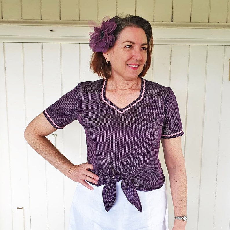 K&ouml;nnte beinhalten: Ein Oberteil mit wei&szlig;en und violetten Punkten und einem rosa Besatz an Ausschnitt und &Auml;rmeln. Das Oberteil ist in der Taille mit einer Schleife gebunden. Das Model tr&auml;gt einen wei&szlig;en Rock.