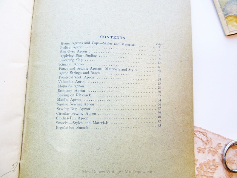 Puede incluir: Un libro vintage con una cubierta de color marr&oacute;n claro y texto azul. El t&iacute;tulo del libro es "CONTENTS" y enumera diferentes tipos de delantales y sus n&uacute;meros de p&aacute;gina correspondientes. El libro se titula "House Aprons and Caps-Styles and Materials" y "Fancy and Sewing Aprons-Materials and Styles".
