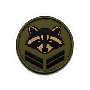 May include: A circular embroidered patch featuring an olive green background with a central raccoon head graphic in black and tan. Below the raccoon head are two parallel black chevron stripes. The patch has a thick olive green stitched border.