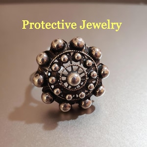 Puede incluir: Anillo plateado con un diseño circular detallado, con una esfera central rodeada de esferas más pequeñas y filigrana intrincada. La inscripción "Protective Jewelry" es visible.