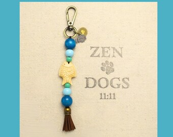 May include: A beaded keychain with a gold-colored clasp, featuring blue, turquoise, and cream-colored beads. A carved fish charm and a small brown tassel hang from the bottom. The background has the text "ZEN DOGS 11:11".