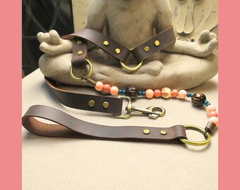 May include: A brown leather harness and leash set with gold-colored hardware. The leash features a handle and a clasp. A beaded strand with coral-colored and brown beads is attached to the leash. The harness has a decorative design.