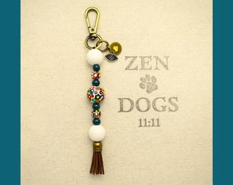 May include: A beaded keychain with a gold-colored clasp and a brown tassel. The keychain features white and colorful beads, along with a small charm with an eye design. The background has the words "ZEN DOGS 11:11" printed on it.