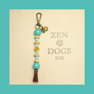May include: A beaded keychain with a bronze clasp, turquoise beads, and a brown tassel. The keychain has a charm with a heart and an eye design. The background has the words "ZEN DOGS 11:11".