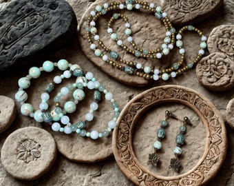 May include: A collection of jewelry featuring a long beaded necklace, three elastic beaded bracelets, and a pair of drop earrings. The pieces incorporate light green, white, and metallic gold beads, along with small turquoise-colored Buddha charms.