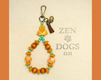 May include: A beaded keychain with a gold-colored frog charm, brown tassel, and various orange, turquoise, and wooden beads. The background features the text "ZEN DOGS 11:11" in a light gray color.