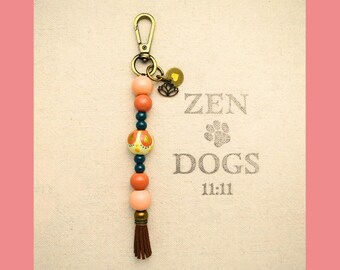 May include: A beaded keychain with a bronze clasp and a brown tassel. The beads are peach, orange, and teal, with a decorative yellow and orange bead. The keychain has a small charm with a lotus flower design. The background has the text "ZEN DOGS 11:11".