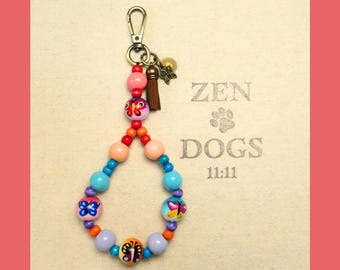 May include: A colorful beaded keychain with a butterfly design. The keychain is attached to a key ring with a small bell charm. The beads are pink, orange, blue, and purple. The keychain has the text "ZEN DOGS 11:11" on it.