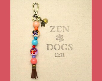 May include: A colorful beaded keychain with a butterfly charm and a tassel. The beads are orange, blue, red, and purple. The keychain has a metal ring and a metal clasp. The text "ZEN DOGS 11:11" is printed on a beige background.