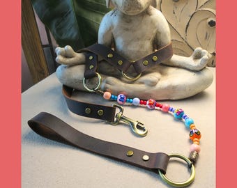 May include: A brown leather restraint with brass hardware and a beaded accent. The restraint features a collar, straps, and a leash with a clasp. The beaded accent includes colorful beads. The items are displayed on a light-colored surface.