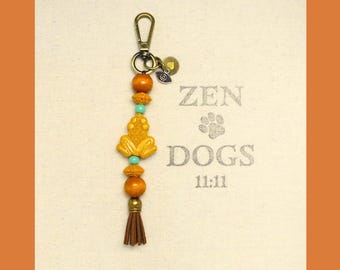 May include: A beaded keychain with a bronze clasp, wooden beads, turquoise accents, and a gold-colored charm. The keychain has a brown tassel and a gold-colored charm with an eye design. The background has the words "ZEN DOGS 11:11".