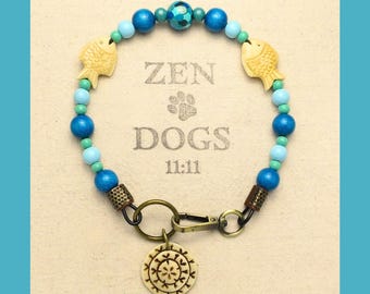May include: A blue and green beaded dog collar with a gold clasp and a wooden charm with a floral design. The collar has two wooden fish charms and the text "ZEN DOGS 11:11" printed on the background.