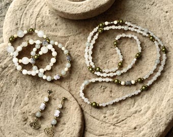 May include: A jewelry set consisting of a long beaded necklace, a multi-strand beaded bracelet, and a pair of drop earrings. The pieces feature white and pale green beads, metallic olive green beads, and silver-toned charms shaped like lotus flowers.