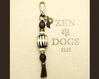 May include: A beaded keychain with a gold-colored clasp and a brown tassel. The keychain features various beads in white, brown, and black, along with a leaf charm. The background has the text "ZEN DOGS 11:11".