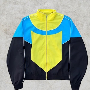 Può includere: Giacca a blocchi di colore giallo, blu e nero. La giacca ha un collo alto, una cerniera a tutta lunghezza e finiture bianche. Le maniche sono nere e il corpo presenta un design geometrico giallo e blu. Un capo di abbigliamento casual.