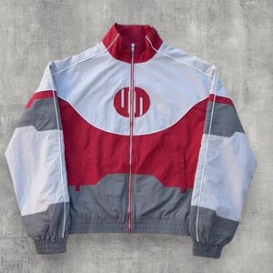 Puede incluir: Chaqueta deportiva roja, gris y blanca con cremallera completa y cuello alzado. La chaqueta presenta un logotipo circular en el pecho y ribetes blancos a lo largo de las costuras. La chaqueta está hecha de un material ligero.