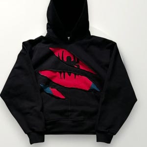 May include: Black hooded sweatshirt with a torn design revealing red and blue accents. The hoodie features a front pocket and a drawstring hood. The design gives the impression of a ripped or torn fabric, with a graphic element visible.