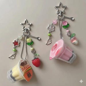 May include: Two keychains with charms. One keychain has a yellow dessert cup, a red strawberry, and a star charm. The other keychain has a pink dessert cup, a strawberry charm, and a flower charm. Both have silver chains and charms.