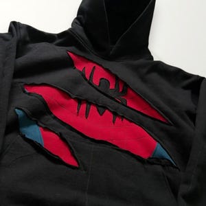 May include: Black hooded sweatshirt with red claw marks revealing a red and blue design. The sweatshirt has a front pocket and a hood. The design appears to be a superhero theme.