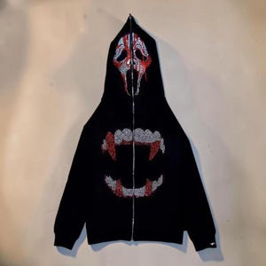 May include: Black zip-up hoodie with a rhinestone design. The hood features a red and silver skull design. The front of the hoodie has a rhinestone vampire teeth design. The hoodie has a silver zipper.