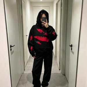 May include: Black hoodie and matching pants. The hoodie features a red design on the front and sleeves. The person in the image is wearing a black hoodie and black pants. The outfit is casual and comfortable.