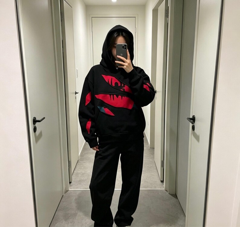 May include: Black hoodie and matching pants. The hoodie features a red design on the front and sleeves. The person in the image is wearing a black hoodie and black pants. The outfit is casual and comfortable.