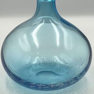 May include: A round, light blue glass bottle with a narrow neck and a rounded top. The bottle has a crackled texture on the lower half and a smooth, glossy finish. The bottle is empty and appears to be a decorative vase.