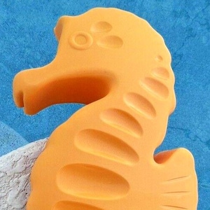 May include: An orange seahorse-shaped ornament with a textured finish. The seahorse features a curled tail and detailed fins. The background is a mix of blue and white, evoking a marine theme.