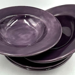May include: A set of purple ceramic plates and bowls. The plates have a broad rim and a shallow centre. The bowls have a deep centre and a wide rim. The plates and bowls are a solid, muted purple colour.