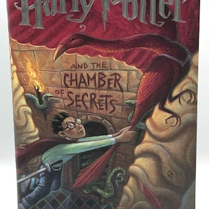 May include: A hardcover book titled Harry Potter and the Chamber of Secrets by J.K. Rowling. The cover illustration depicts a young person with glasses and a sword reaching toward a large red bird in a stone chamber with green snakes and a torch.