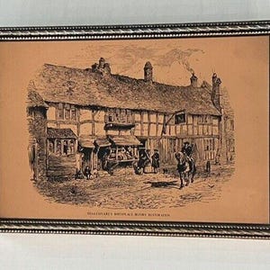May include: Framed black and white etching of Shakespeare's birthplace before restoration. The artwork depicts a detailed street scene with a large building, figures, and a horse-drawn carriage. The frame is silver.