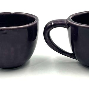 May include: Two dark purple ceramic mugs with curved handles. The mugs have a smooth, glossy finish and a rounded shape. The interior of the mugs is also dark purple.