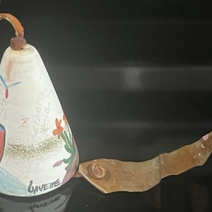 May include: A decorative bell with a white conical body, adorned with colorful illustrations of birds and flowers. It features a brown leather handle and a long, curved metal clapper. The bell is signed "Gavette".