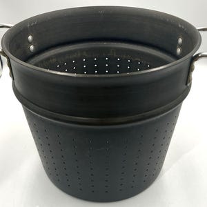 Può includere: Uno scolapasta in metallo grigio scuro con un bordo circolare ampio e un corpo affusolato dotato di numerosi piccoli fori di drenaggio. L'utensile da cucina include due manici curvi in metallo color argento fissati ai lati con rivetti.