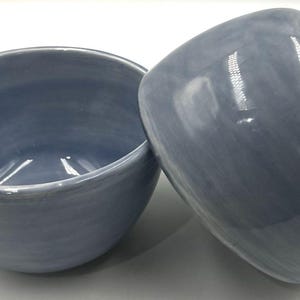 May include: Two ceramic bowls with a smooth, glossy finish in a muted periwinkle blue color. One bowl is positioned upright to show the interior, while the second bowl is tilted on its side, revealing the rounded exterior and the white base rim.