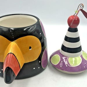 May include: A whimsical ceramic container and lid set. The container is black with a toucan face design in orange, red, and yellow. The lid is purple with green polka dots and a black and white striped cone topped with a red sphere and maroon wings.