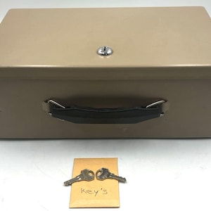 May include: A rectangular beige metal cash box with a black plastic handle and a silver key lock on the top lid. Two silver metal keys are placed on a small tan paper envelope with the handwritten word Key's visible on the front surface.