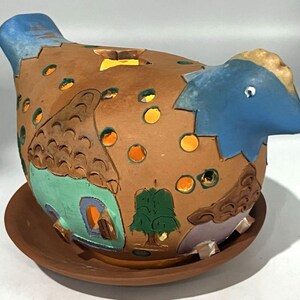 May include: A terracotta chicken-shaped luminary featuring a blue head and tail, with cut-out star and circular patterns. The body is decorated with painted houses, a green tree, and a light purple structure, all resting on a matching terracotta saucer.