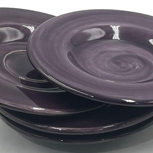 Puede incluir: Una pila de cuatro platos redondos de cerámica en un tono morado intenso. Cada plato presenta un acabado liso y brillante con sutiles texturas de pinceladas en espiral visibles en toda la superficie, creando un aspecto uniforme.