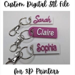 Op de afbeelding: Drie witte en roze sleutelhangers met de namen "Sarah", "Claire" en "Sophia" erop gedrukt. De sleutelhangers zijn voor 3D-printers.