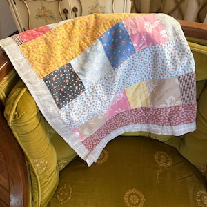May include: A patchwork quilt featuring a variety of fabric squares in yellow, pink, blue, brown, and white. The quilt displays diverse patterns including floral prints, gingham checks, and solid colors, finished with a white border along the edges.