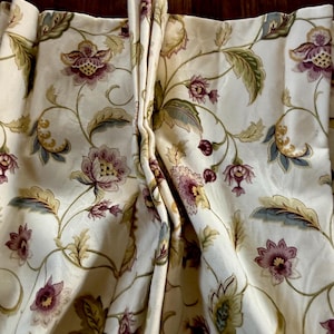 May include: Cream-colored floral curtains with a repeating pattern of burgundy and purple flowers, green leaves, and blue accents. The fabric is gathered at the top, creating soft folds. The curtains have a classic, elegant design.