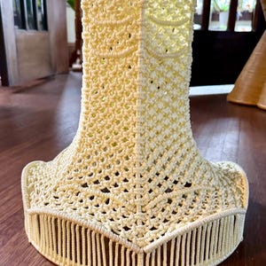 May include: Cream-colored macrame lampshade with a flared base and a cylindrical top. The lampshade features a geometric open weave design, with vertical fringe detailing the bottom edge. The overall shape is reminiscent of a bell, suitable for a table lamp.