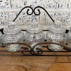 May include: A decorative spice rack with five clear glass jars with lids. The jars have a textured design and sit in a wrought iron rack. The rack has a curved design and is brown.