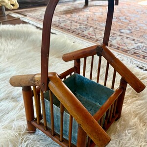 May include: A brown bamboo basket with a tall arched handle and a square metal insert. The structure features vertical bamboo slats forming the sides, with angled bamboo pieces at the top corners, creating a frame around the central metal container.