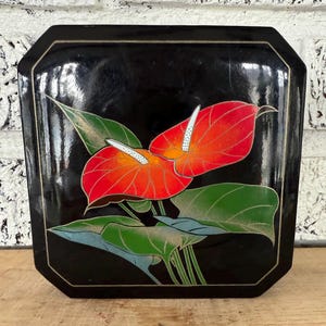May include: A black lacquered wooden box with a square shape and a beveled edge. The lid features a hand-painted design of two red anthurium flowers with green leaves and gold accents.