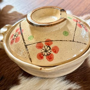May include: A beige ceramic lidded pot with a handle on each side. The lid is decorated with red floral designs and green dots, outlined with black lines. The pot is on a brown and white surface.