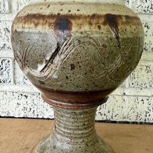 May include: A ceramic chalice with a wide, rounded bowl and a pedestal base. The piece features a speckled green and brown glaze with a smooth, cream-colored rim. The body is decorated with etched, swirling line patterns and dark brown accents.