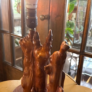 May include: A table lamp featuring a base crafted from three polished, reddish-brown natural wood segments mounted on an oval wooden platform. A single light bulb is attached to the top of the tallest wood segment, emitting a warm glow.
